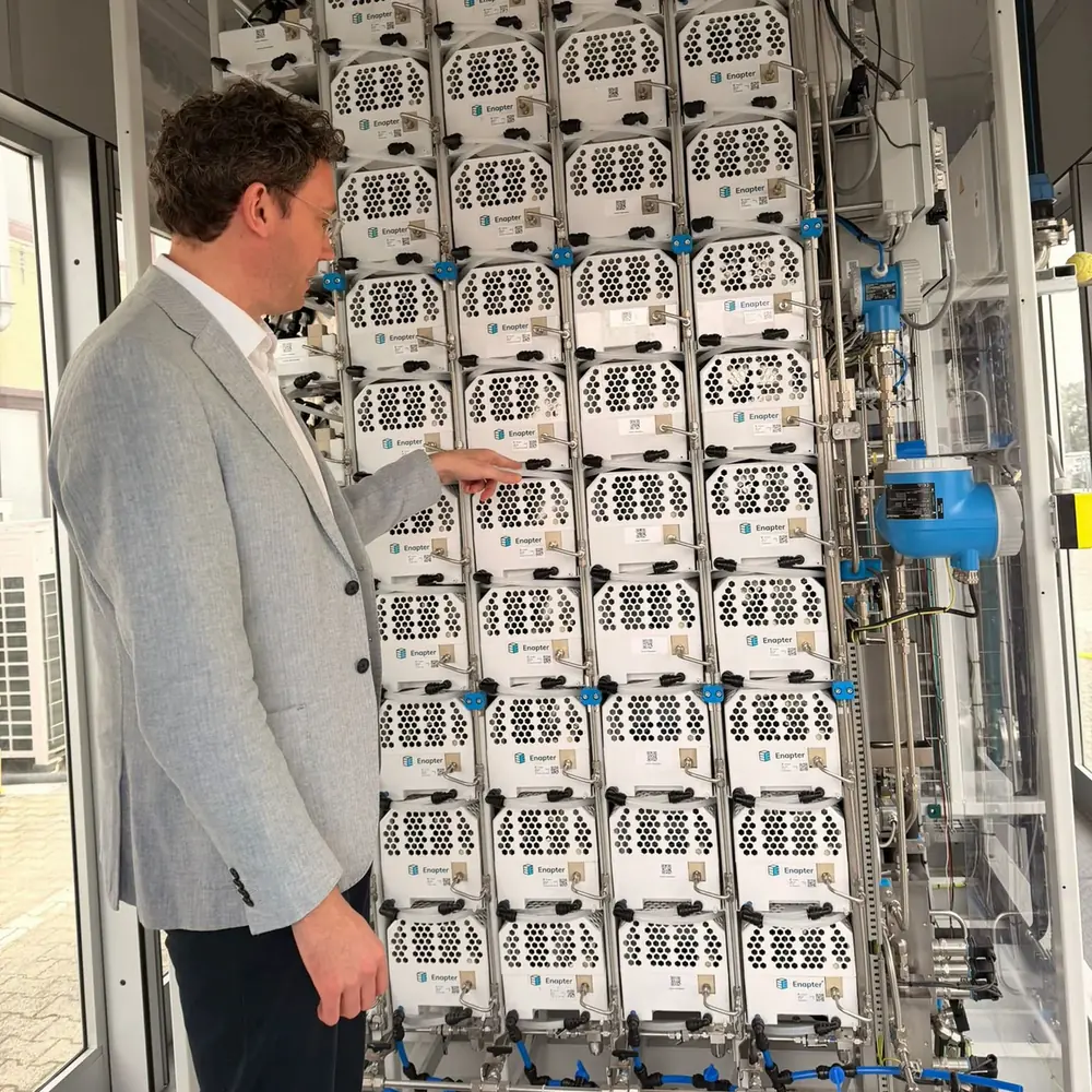 A rack of dozens of identical Enapter AEM electrolyser modules in a containerised plant, with pipework and valves down one side and a person at it for scale